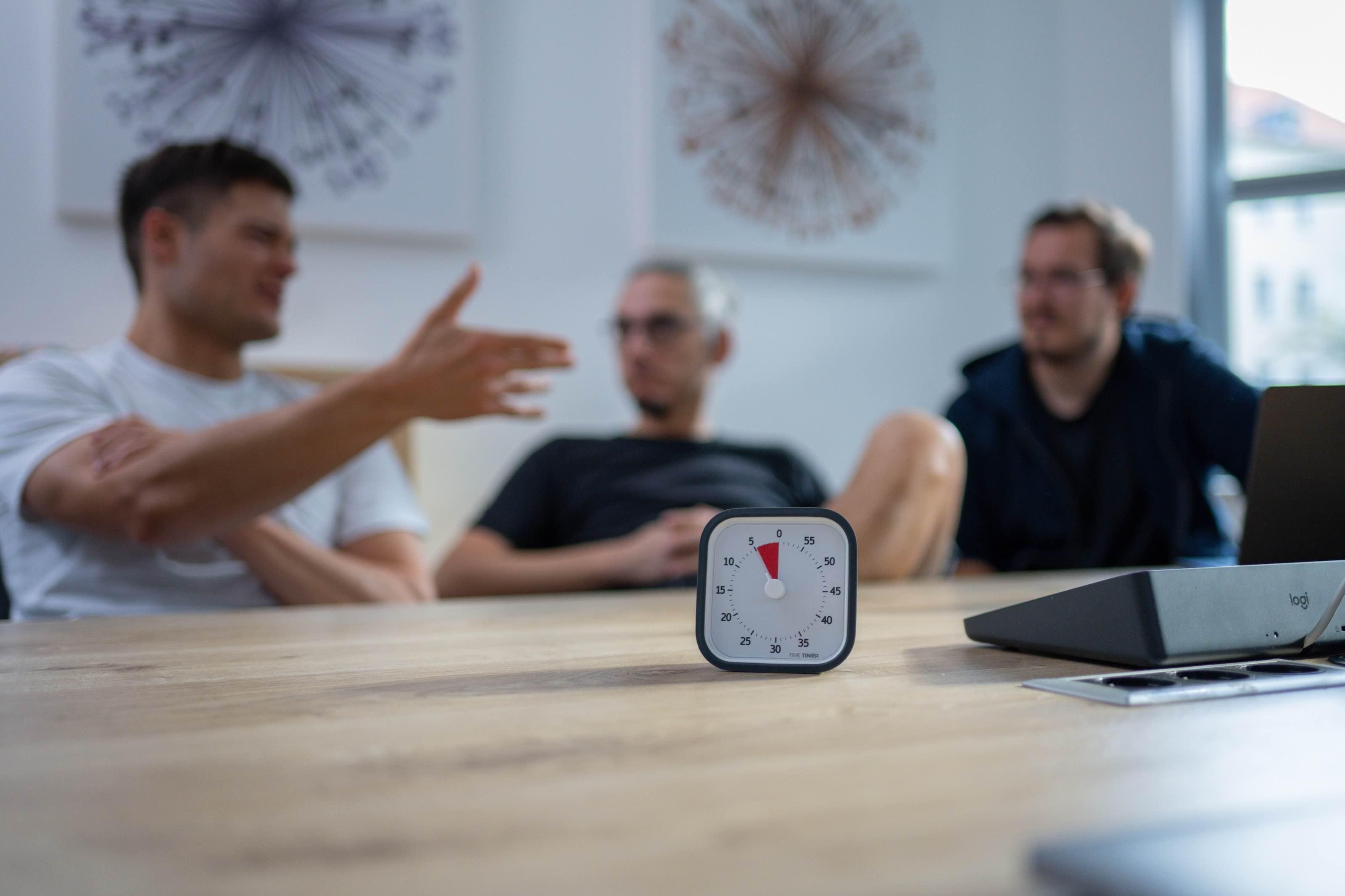 Drei Kollegen diskutieren an einem Tisch; vor ihnen steht ein Timer.