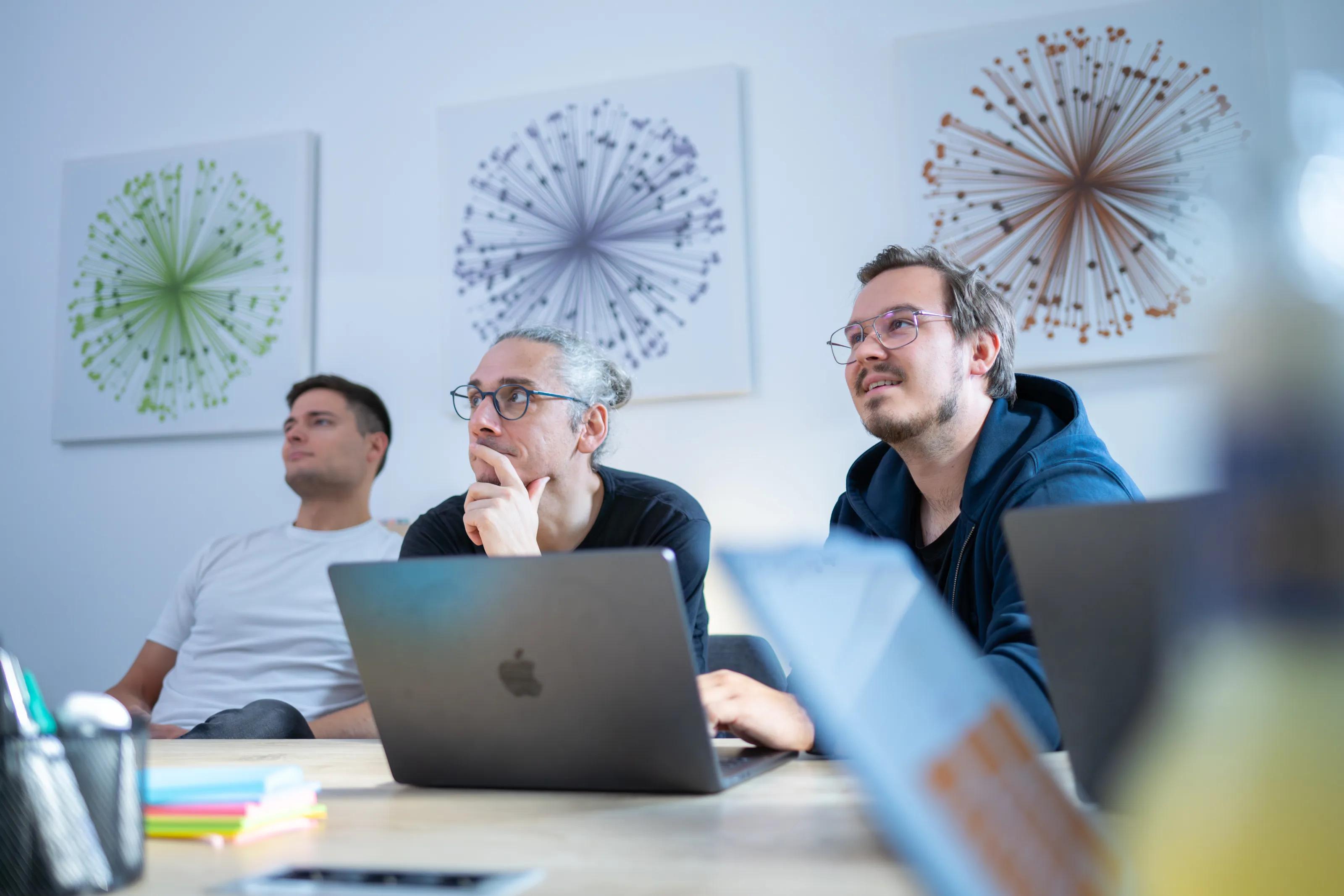 Drei Entwickler schauen gemeinsam auf den Bildschirm eines MacBooks und arbeiten an Docker Sandboxes