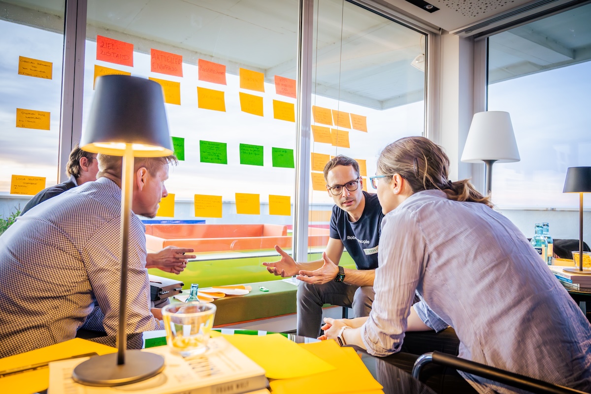 Vier Personen sitzen in einem hellen Meetingraum vor einer Glaswand mit bunten Post-its und diskutieren intensiv über ein Projekt.